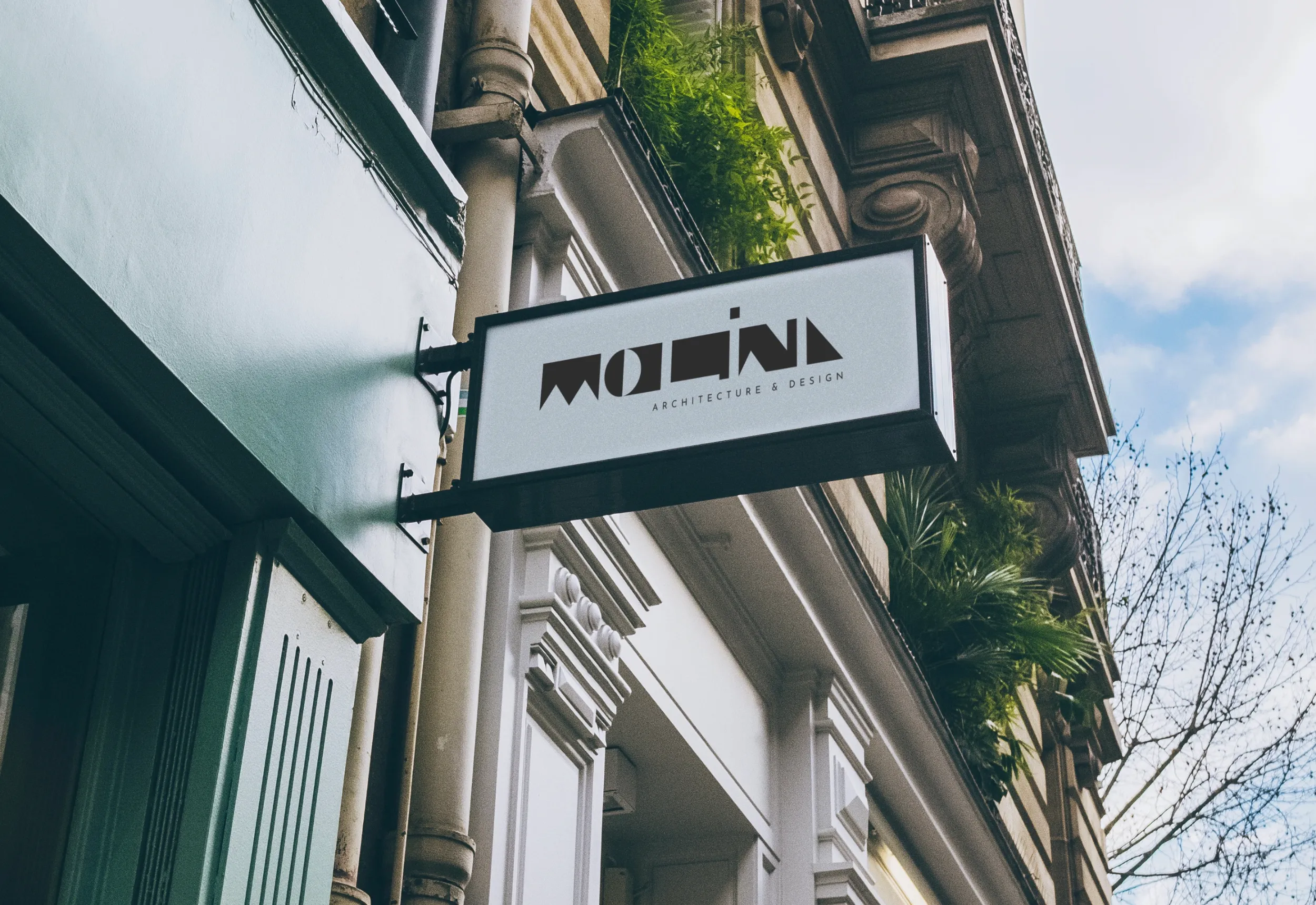 Rectangular sign reading 'MOLINA ARCHITECTURE & DESIGN' mounted on the side of a building with ornate white trim and greenery above.
