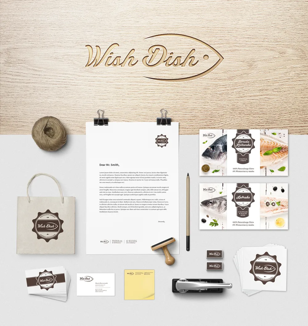 Flat lay of branding materials for Wish Dish including a letterhead, business cards, paper bag, stamps, a pencil, a stapler, and product labels featuring fish images on a wooden and white surface.