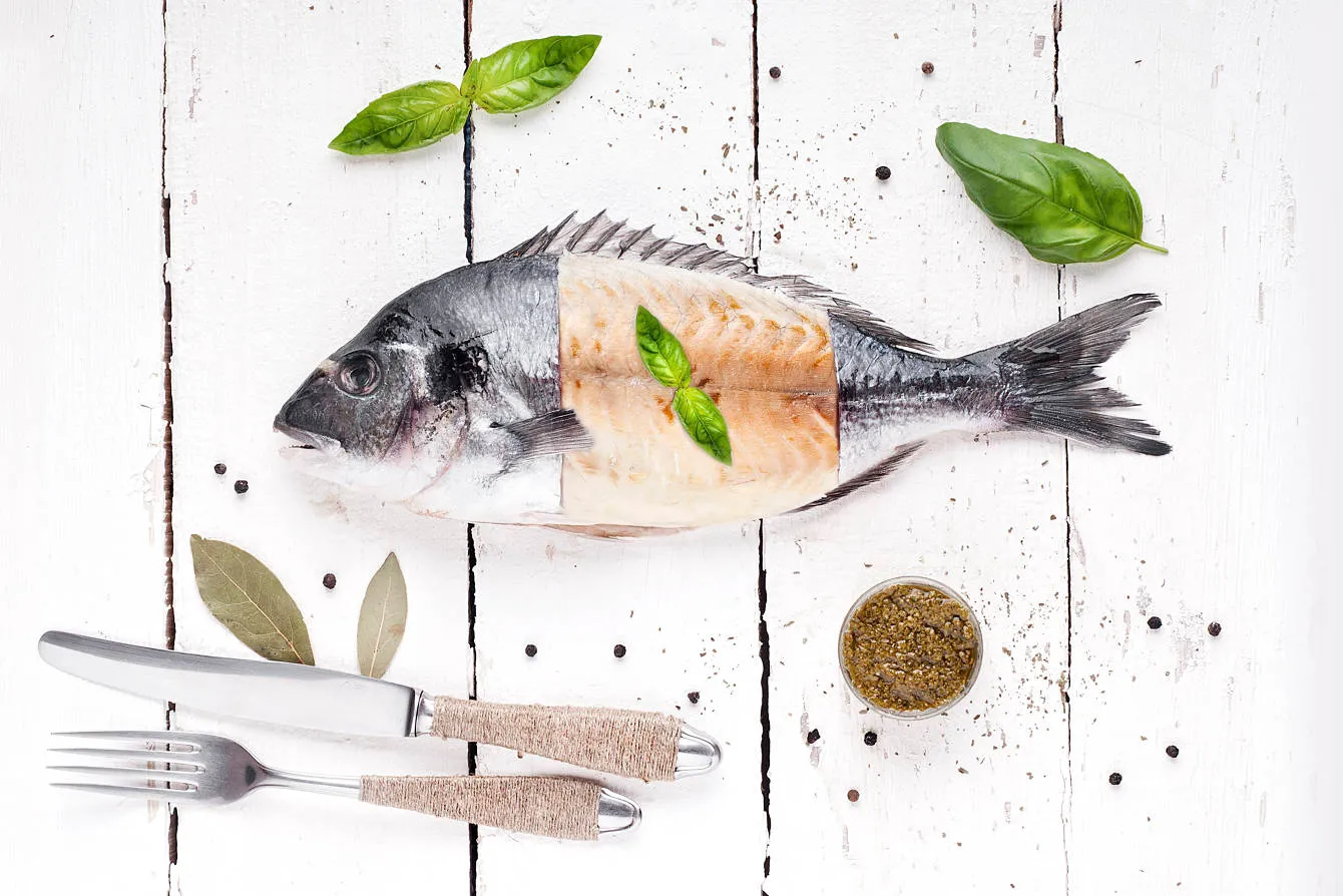 Whole fish with the middle section filleted, garnished with basil leaves, placed on a white wooden surface with scattered peppercorns, bay leaves, and a fork and knife.
