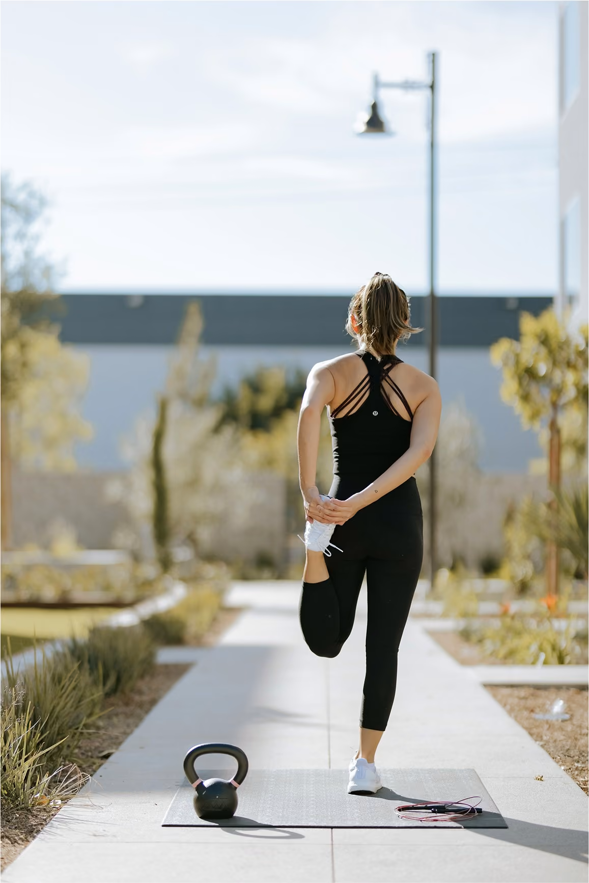 A woman in black activewear stretching on a yoga mat outside with a kettlebell to her left.
