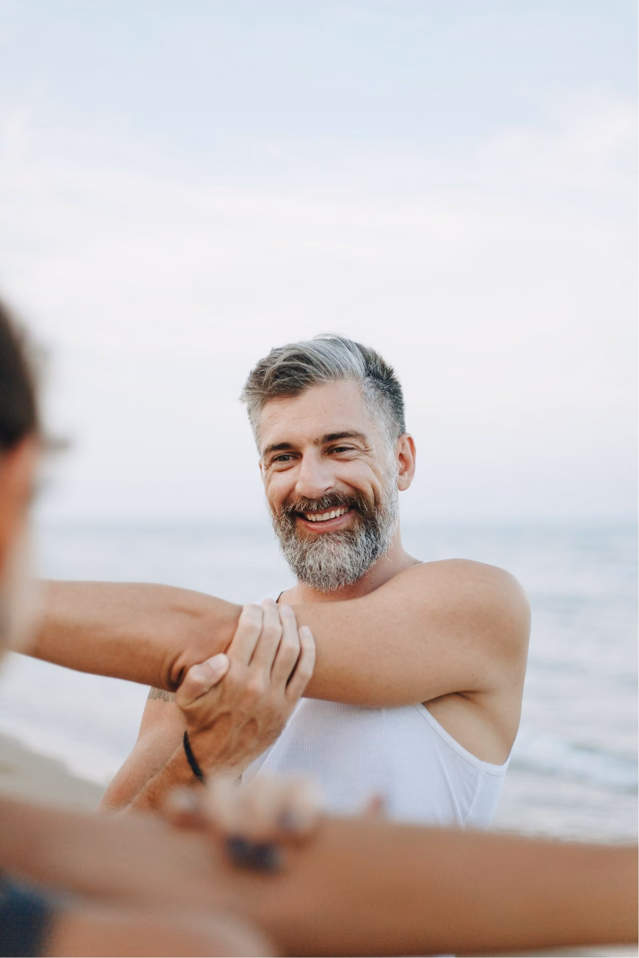 A man in a white singlet stretching his shoulder on the beach
