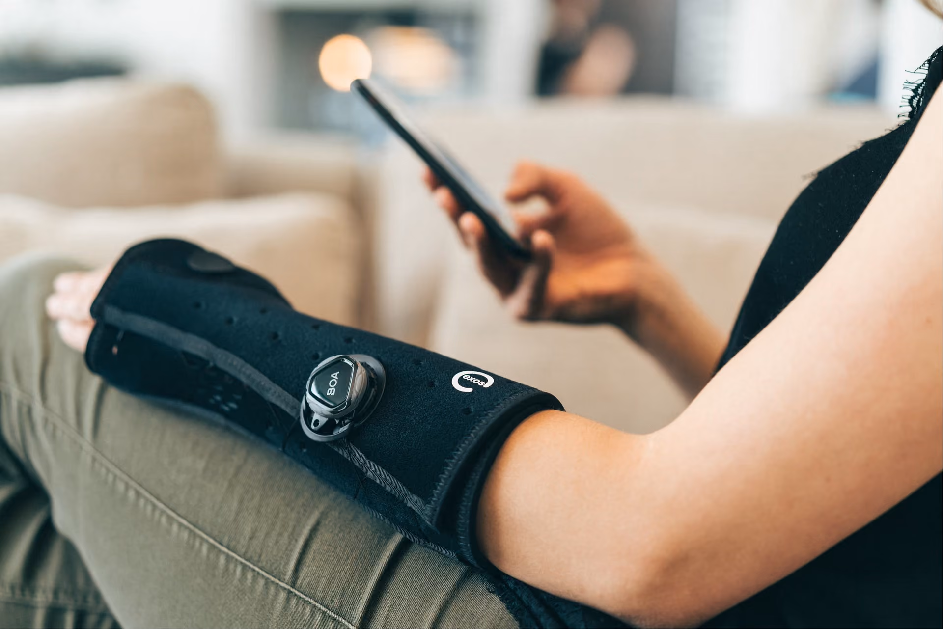 A close-up image of a woman in a black shirt and green pants sitting down, with an EXOS brace on her left wrist.