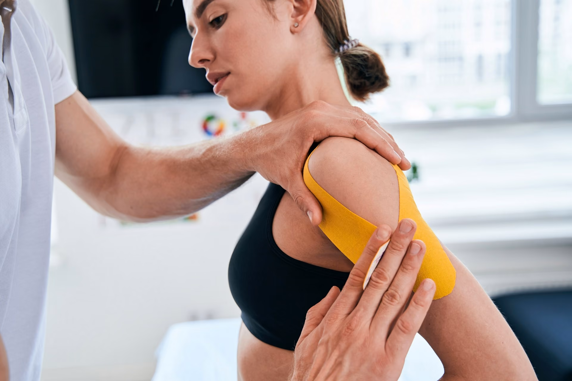 A physiotherapist applying yellow kinesiology tape to a woman's shoulder in a clinic