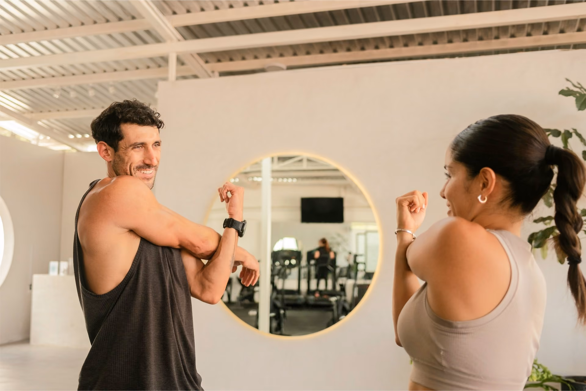 A man in a black single and a woman in a cream-coloured shirt stretching in a fitness studio, with a woman on a treadmill in the reflection of a mirror on the wall.