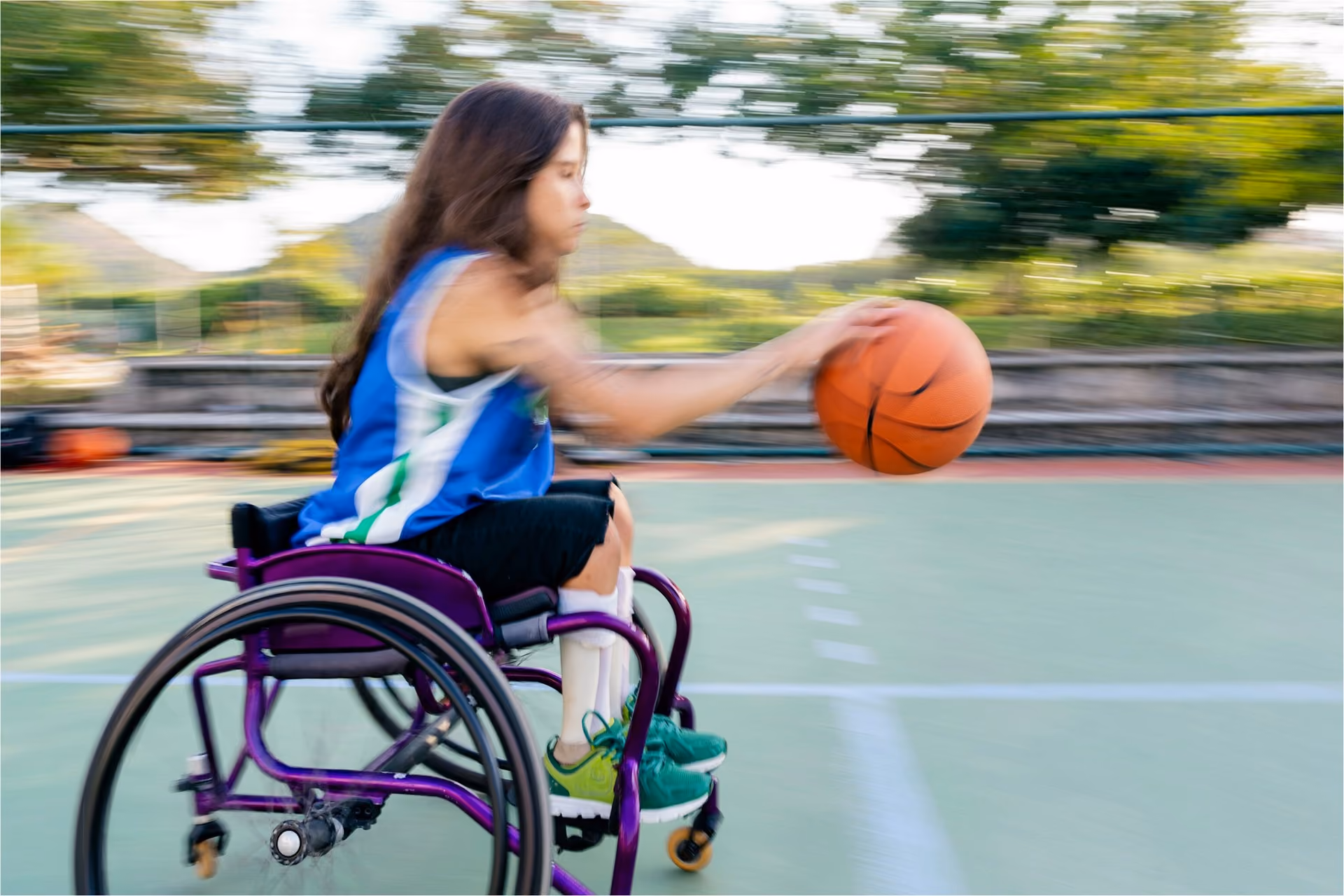 A motion shot of a woman in a blue singlet in a wheelchair playing basketball during the day.