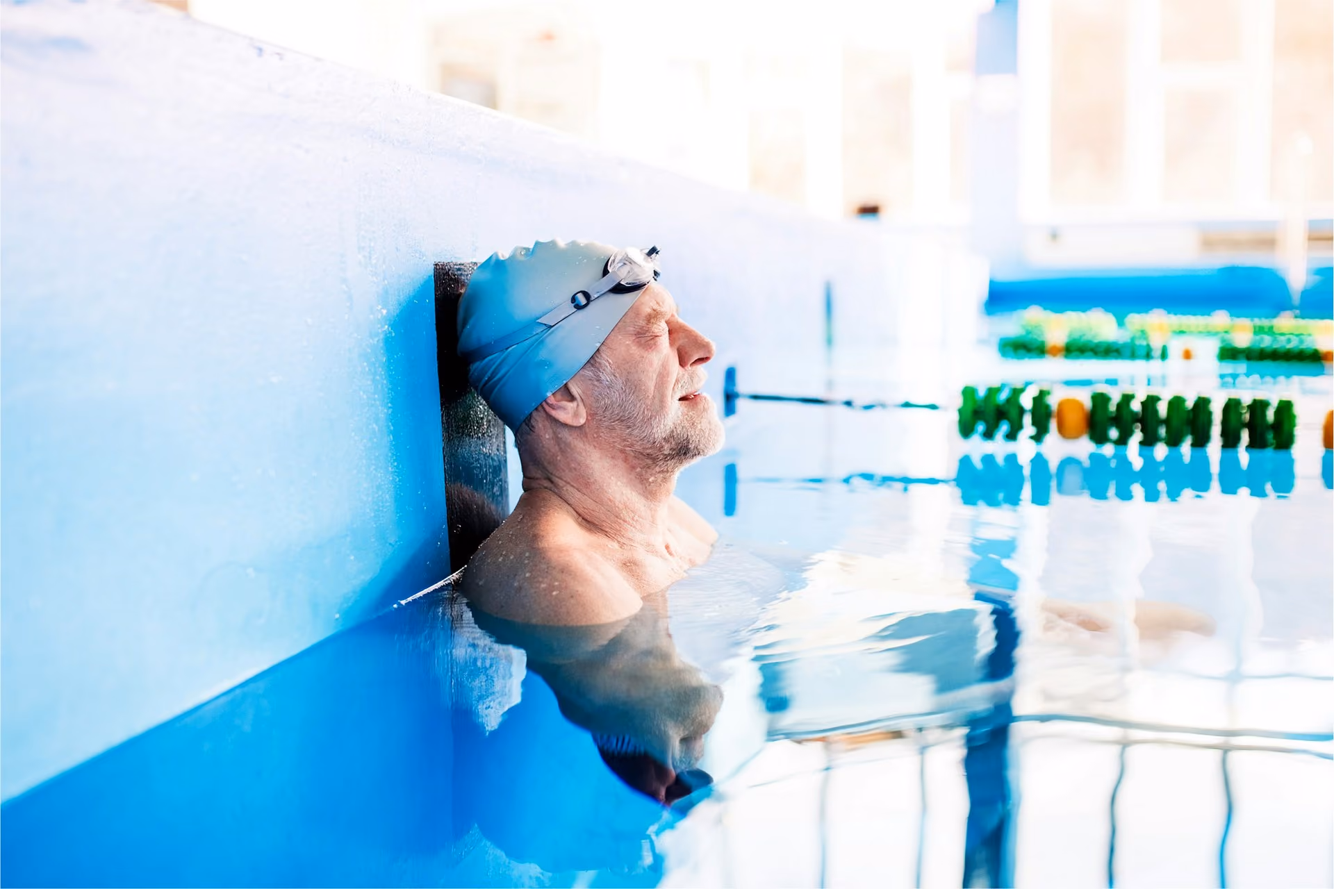 An elderly man cweating a swimming cap and goggles, closing his eyes whilst his back is to the edge of the swimming pool.