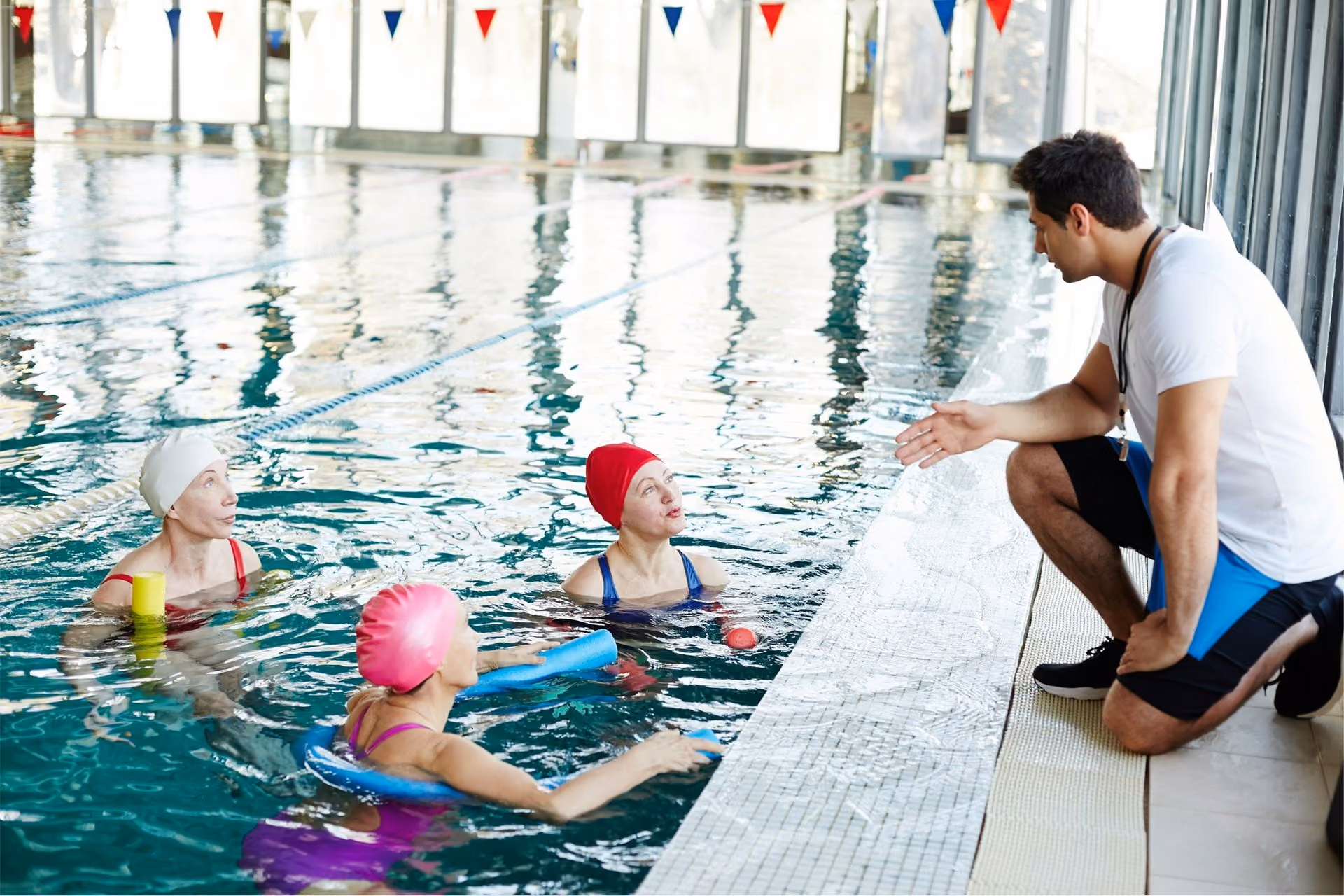 A group of elderly women in a swimming pool with swimming caps on, listening to an instructor who's kneeling at the edge of the pool.
