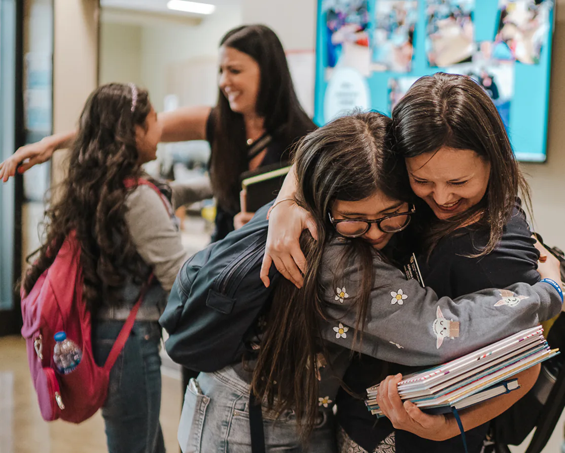 Two girls with backpacks warmly hugging two women inside a school hallway.