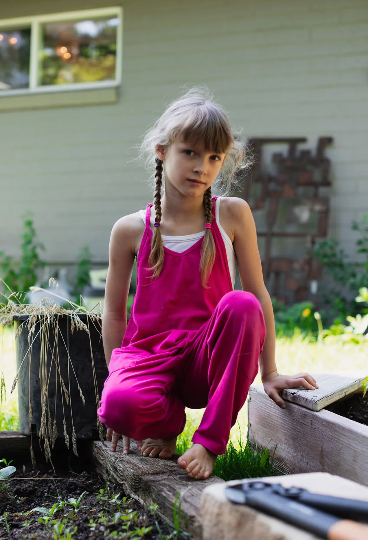 Young girl with braided hair wearing a pink outfit crouching barefoot in a garden near raised wooden garden beds.