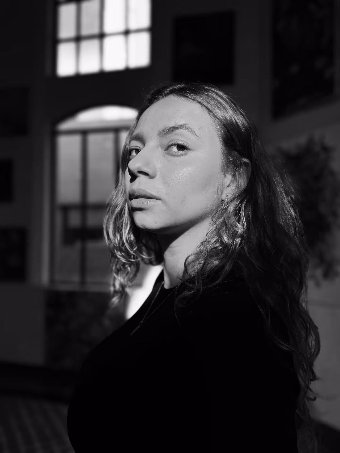 Black and white portrait of a woman with long hair looking sideways in front of a large window.