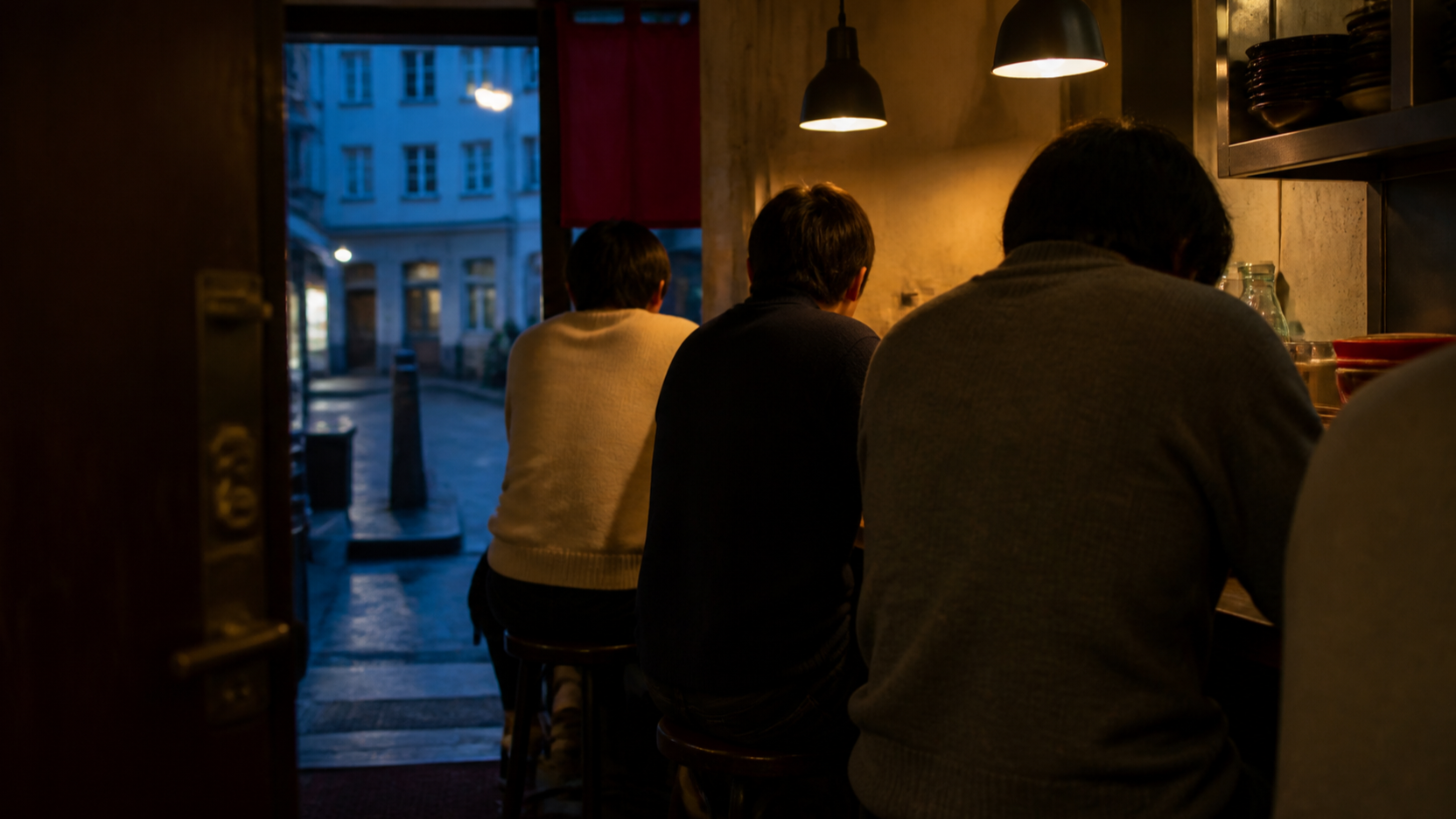 Warm-lit small ramen counter room seen from the entrance at blue hour, diners seated with their backs to camera and a Copenhagen street visible through the open door.