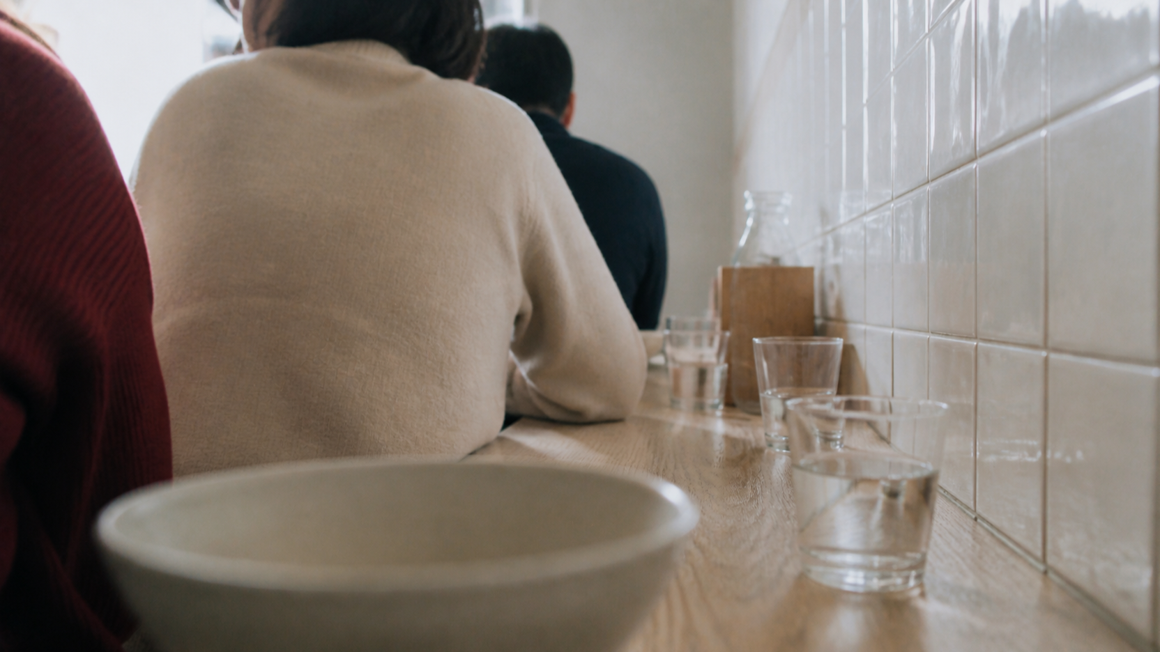 A small counter seen from a seated diner's eye level in daylight, with a soft out-of-focus bowl in the foreground, other diners seated further along with their backs to camera, against a white tiled wall.