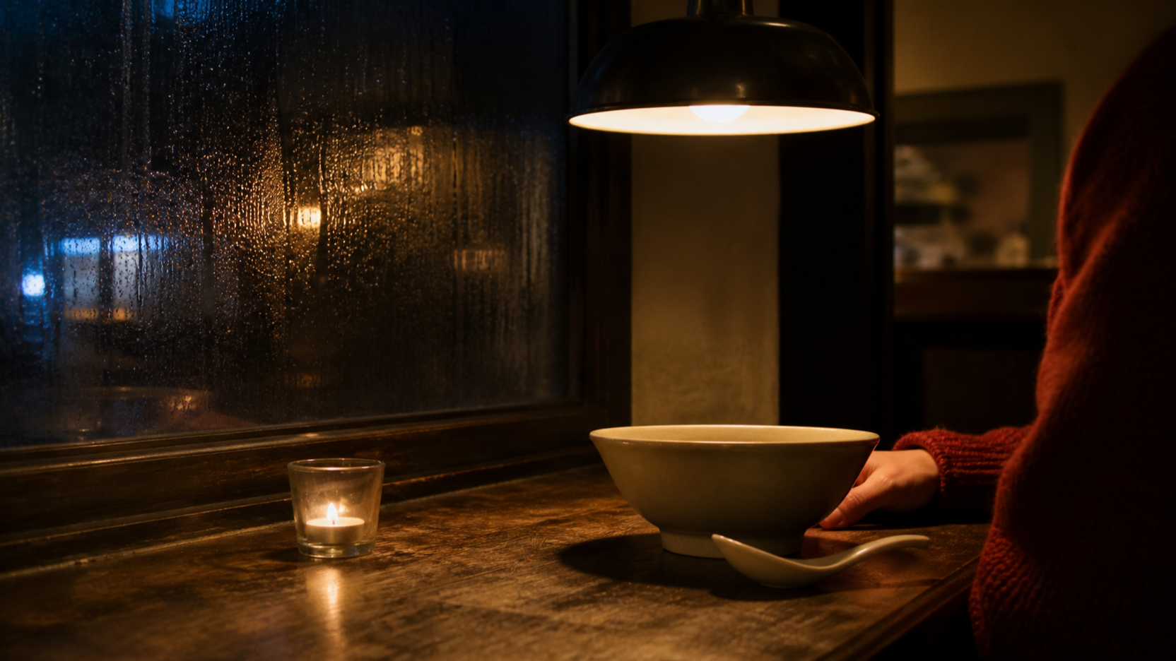 A candlelit restaurant corner with condensation on a dark window, a knitted sleeve and hand resting near a bowl.