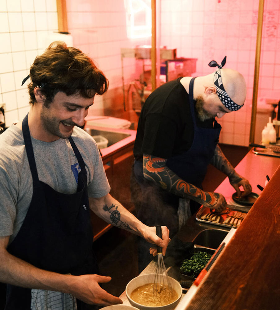 Two Slurp cooks working the pass under warm light, one whisking a small bowl at the counter while the other works the grill behind