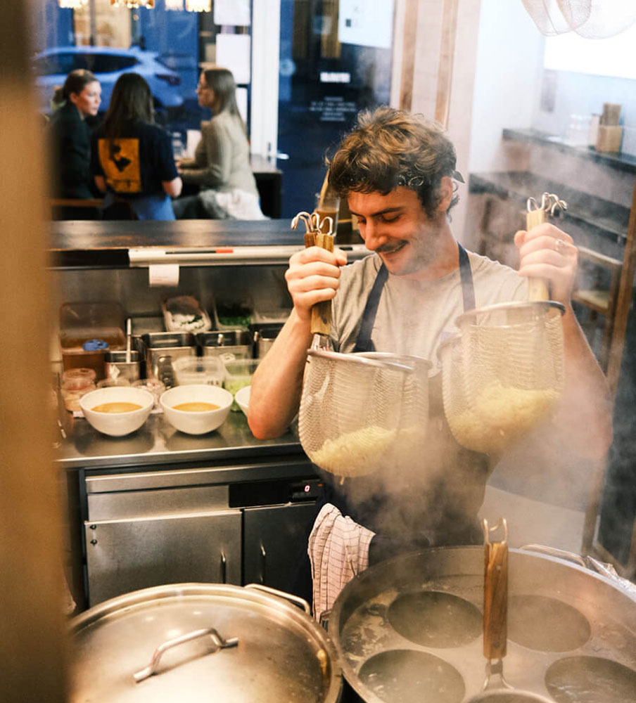 A Slurp cook lifting two noodle baskets out of the steam above the stockpots, with filled bowls waiting on the pass