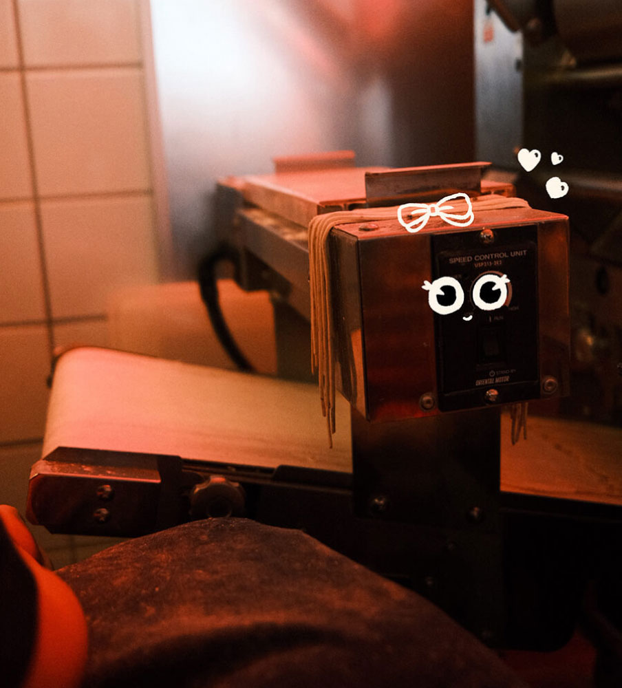 Fresh-cut ramen noodles draped over the noodle machine in the Slurp kitchen, its speed control unit lit warm red against white wall tiles, with Slurp's drawn eyes and bow mark on the housing.