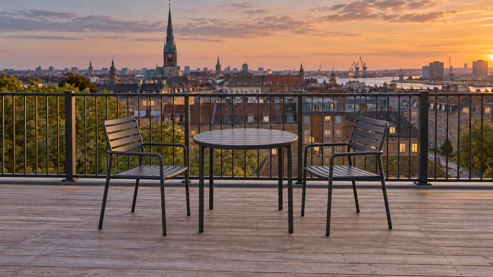 A rooftop terrace above Copenhagen at dusk, a round metal table and two chairs on weathered decking inside the railing, with tiled roofs, a copper spire and harbour cranes going blue beyond