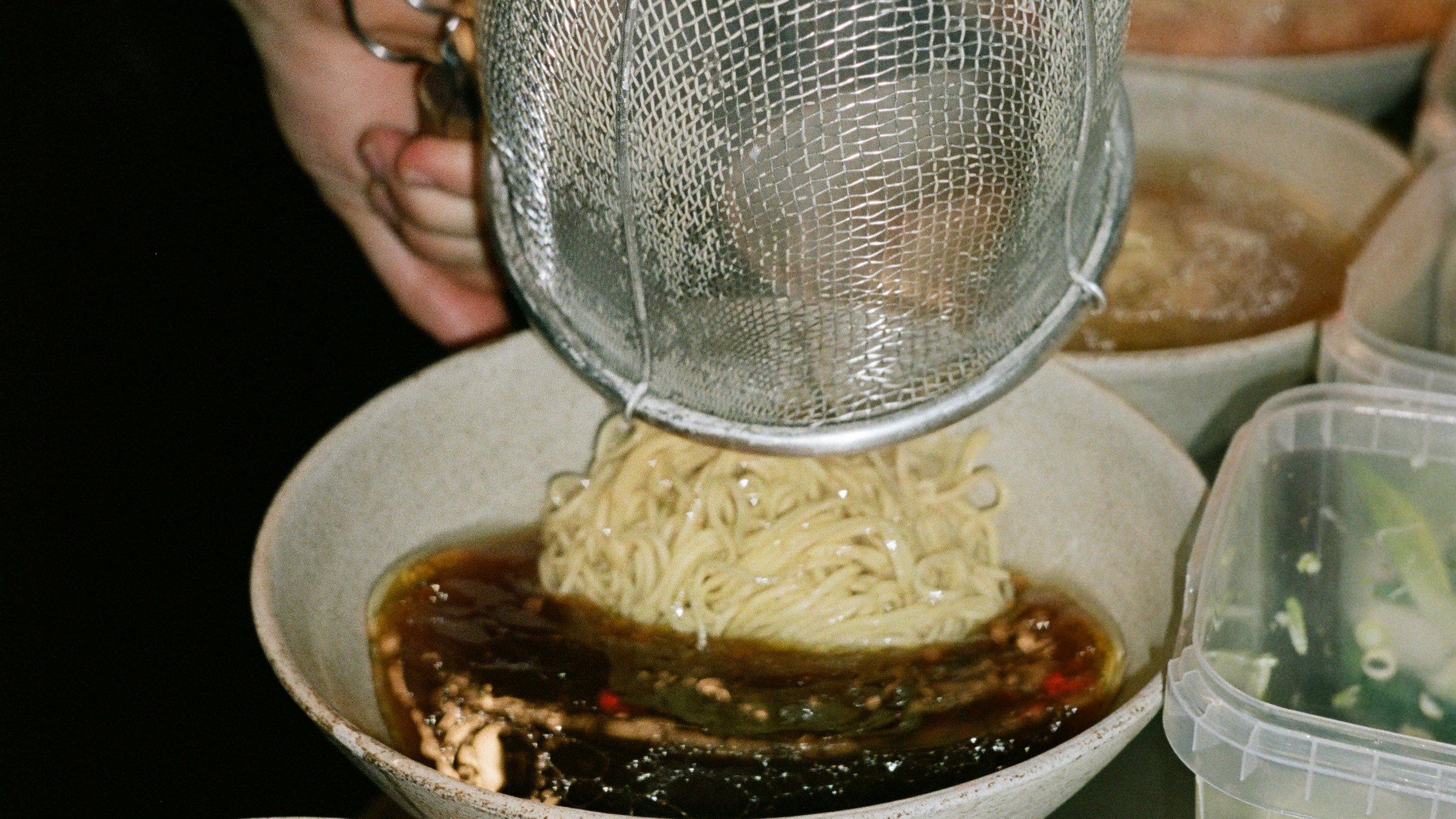 Noodles lifted from a steaming strainer into a finished bowl at Slurp's pass