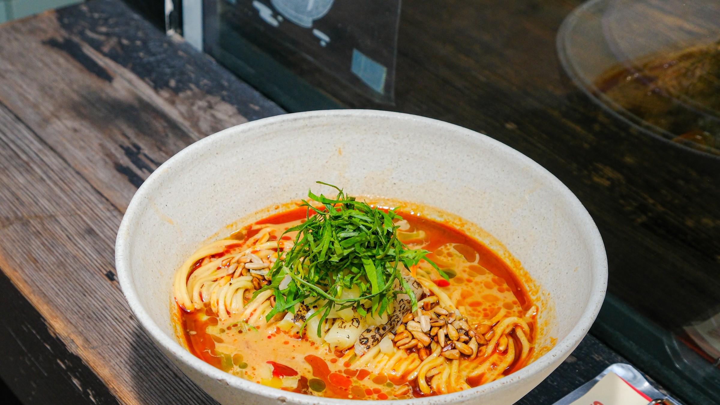 A bowl of ramen topped with shredded shiso and sunflower seeds on a dark wooden counter beside a window