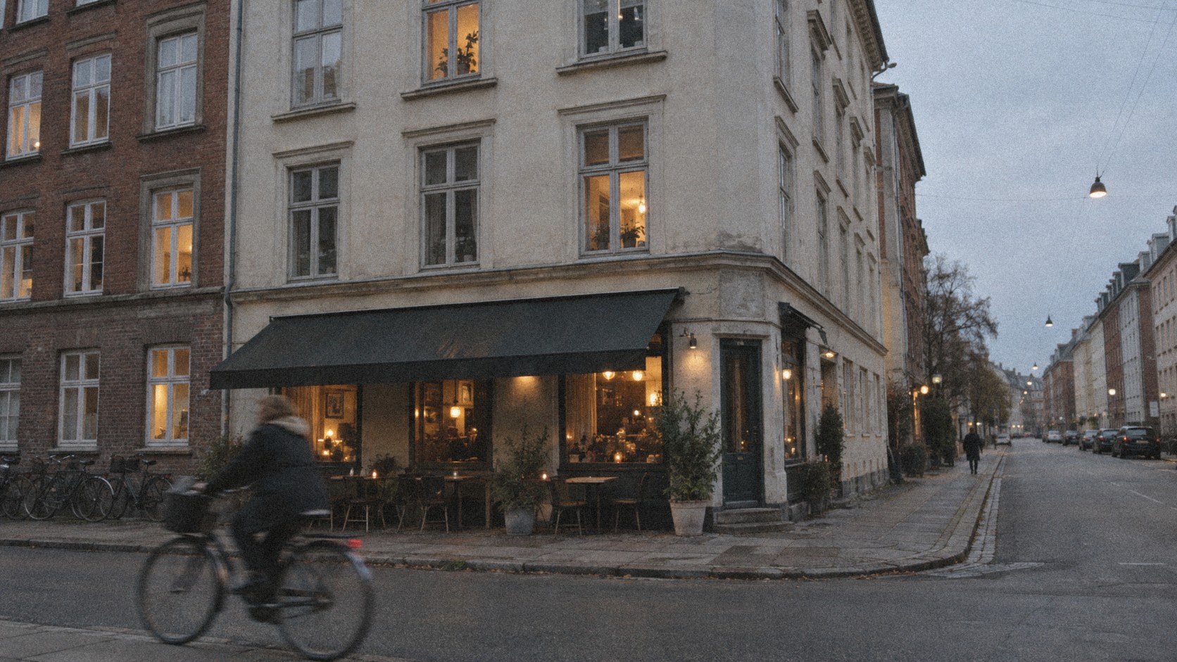 A quiet Copenhagen street corner at dusk with a lit corner cafe and bicycles leaning against the facade