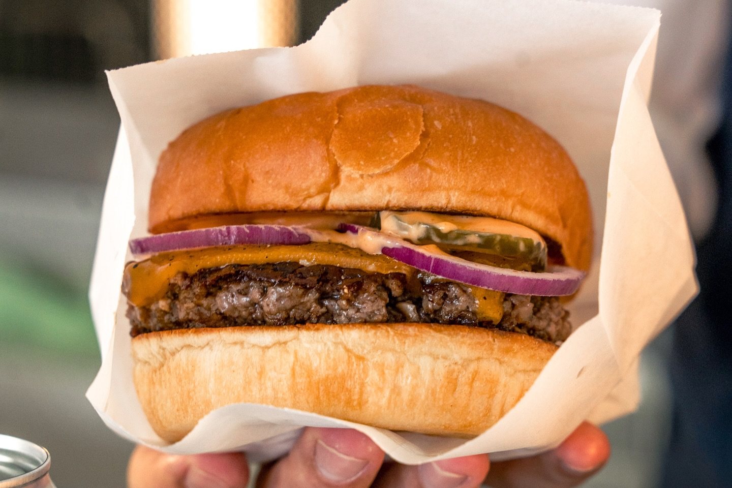 A hand holding a cheeseburger in a white paper wrap, with one patty, melted cheddar, red onion, pickle and sauce on a glossy bun