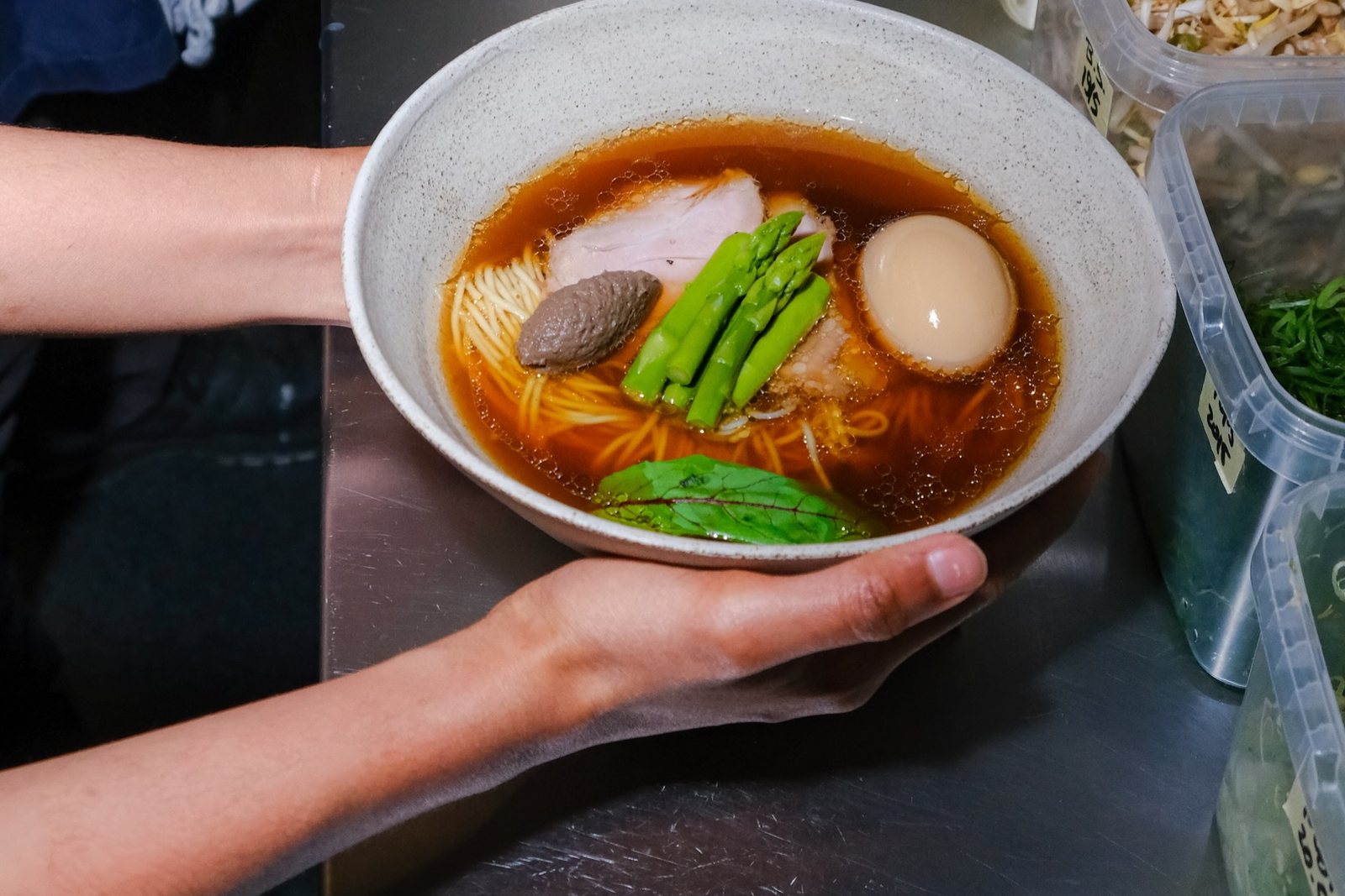 Two hands holding a bowl of shoyu ramen over a steel counter, with a slice of pork chashu, a soft-boiled egg, a dark mushroom paste, green spears and a sorrel leaf in brown broth, and tubs of garnish beside it