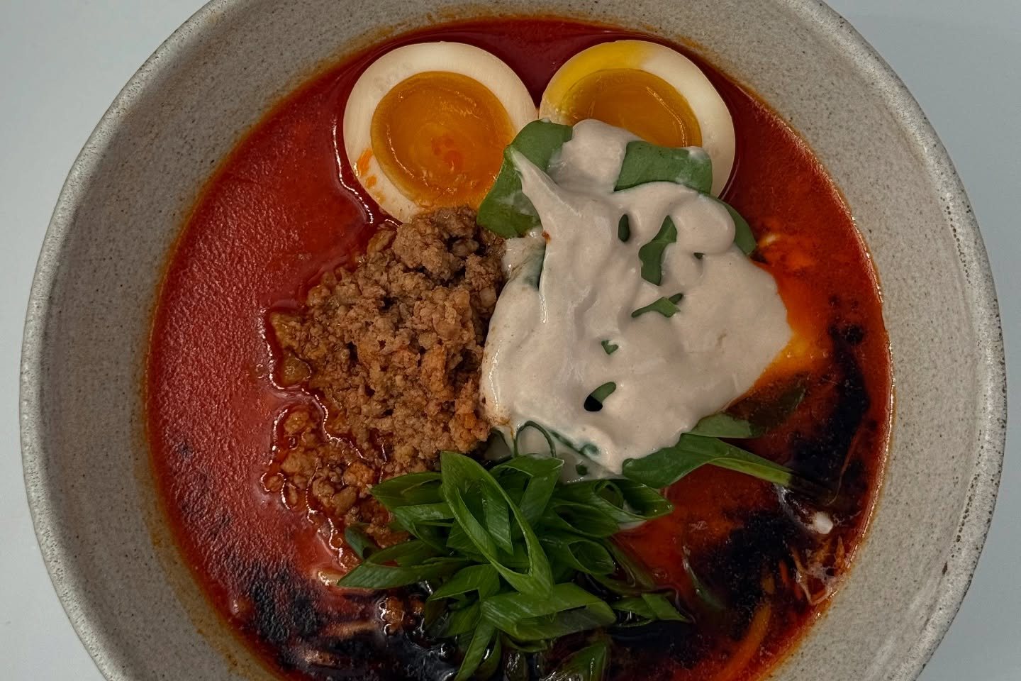 Overhead view of a bowl of red nduja miso ramen with minced pork, two halves of soft-boiled egg, a spoonful of cream and chopped spring onion in a grey stoneware bowl
