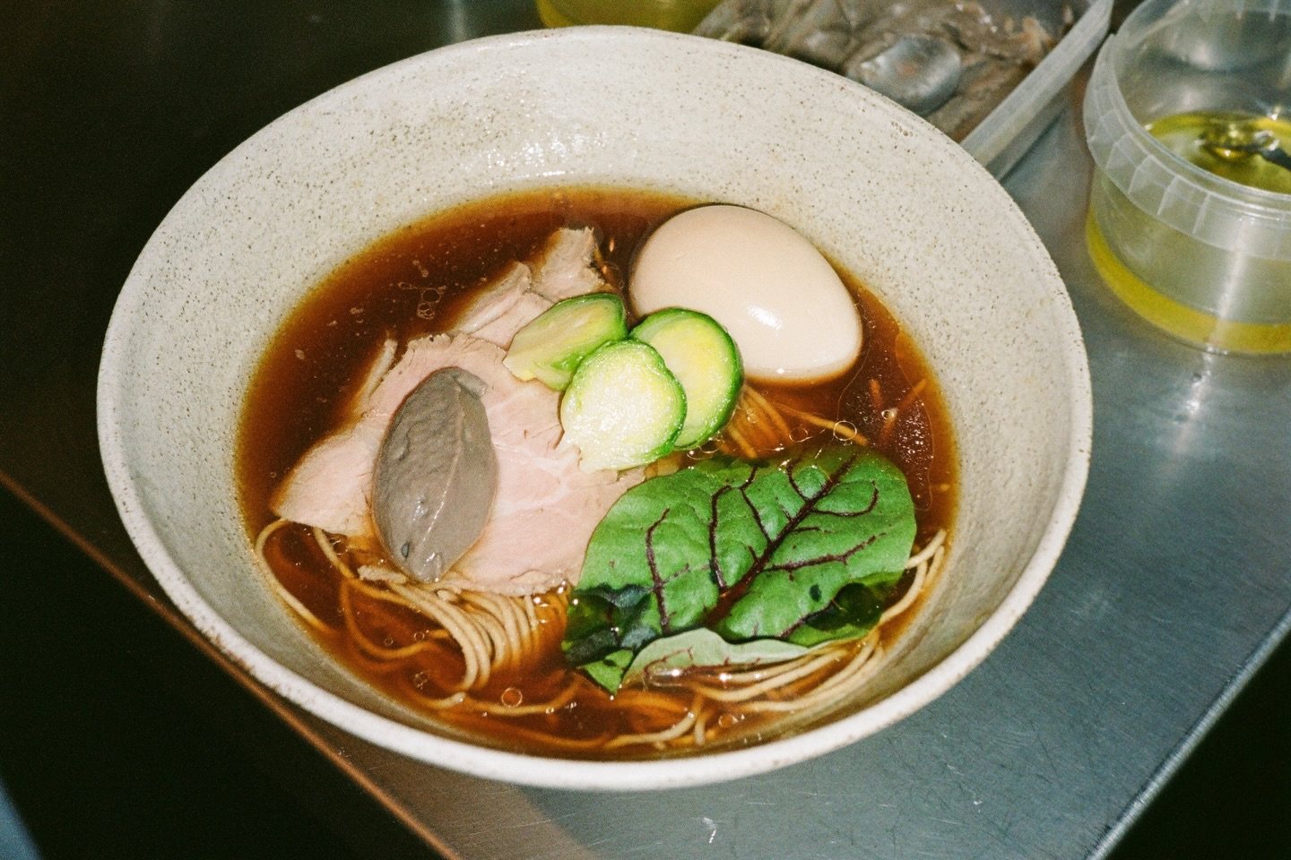 A bowl of shoyu ramen on a steel counter with sliced pork, a soft egg, green leaves and a spoonful of mushroom paste in a dark broth