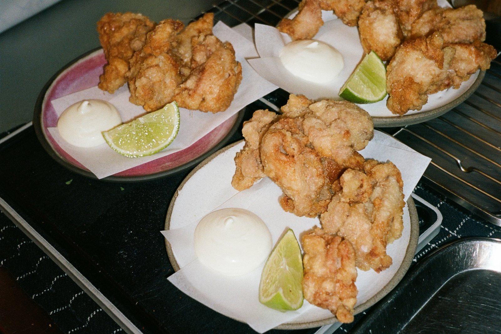 Three plates of fried chicken, each with a swirl of mayonnaise and a lime wedge, on a black tray beside a steel rack