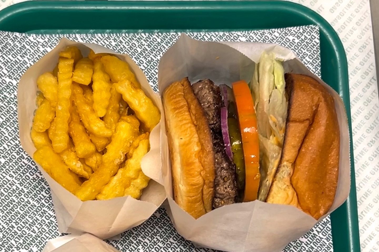 A burger with lettuce, tomato, red onion and pickles in paper, beside a carton of fries on a green tray