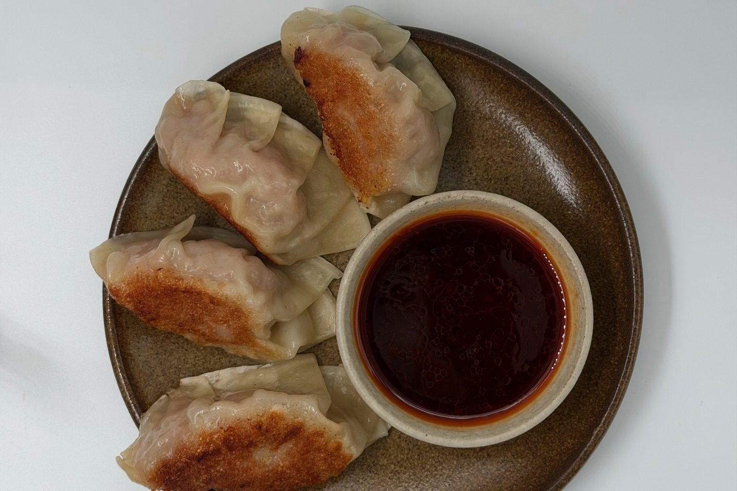 Four pan-fried pork gyoza on a brown plate beside a small bowl of dark dipping sauce