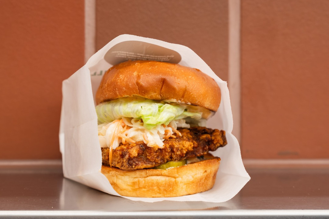 A hot chicken sandwich in white paper with a crisp chicken thigh fillet, coleslaw, lettuce and pickle in a glossy bun, on a steel counter against terracotta tiles