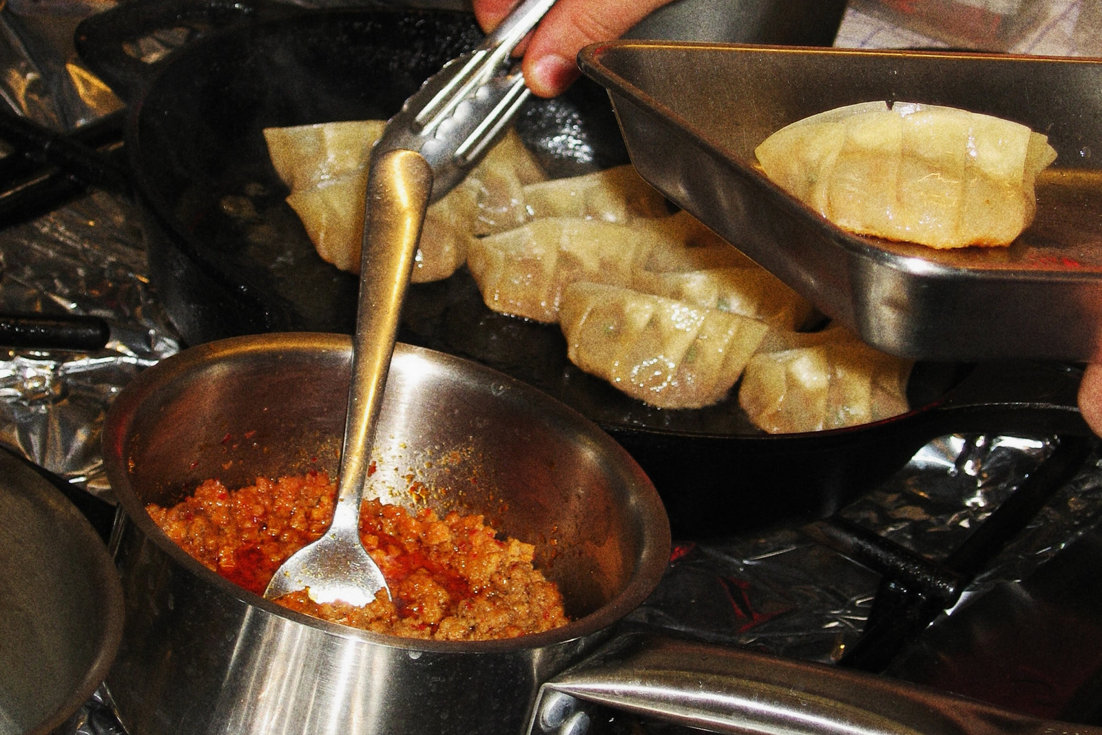 Pan-fried gyoza in a black pan, with a hand lifting one out with tongs into a steel tray beside a pot of chilli mince