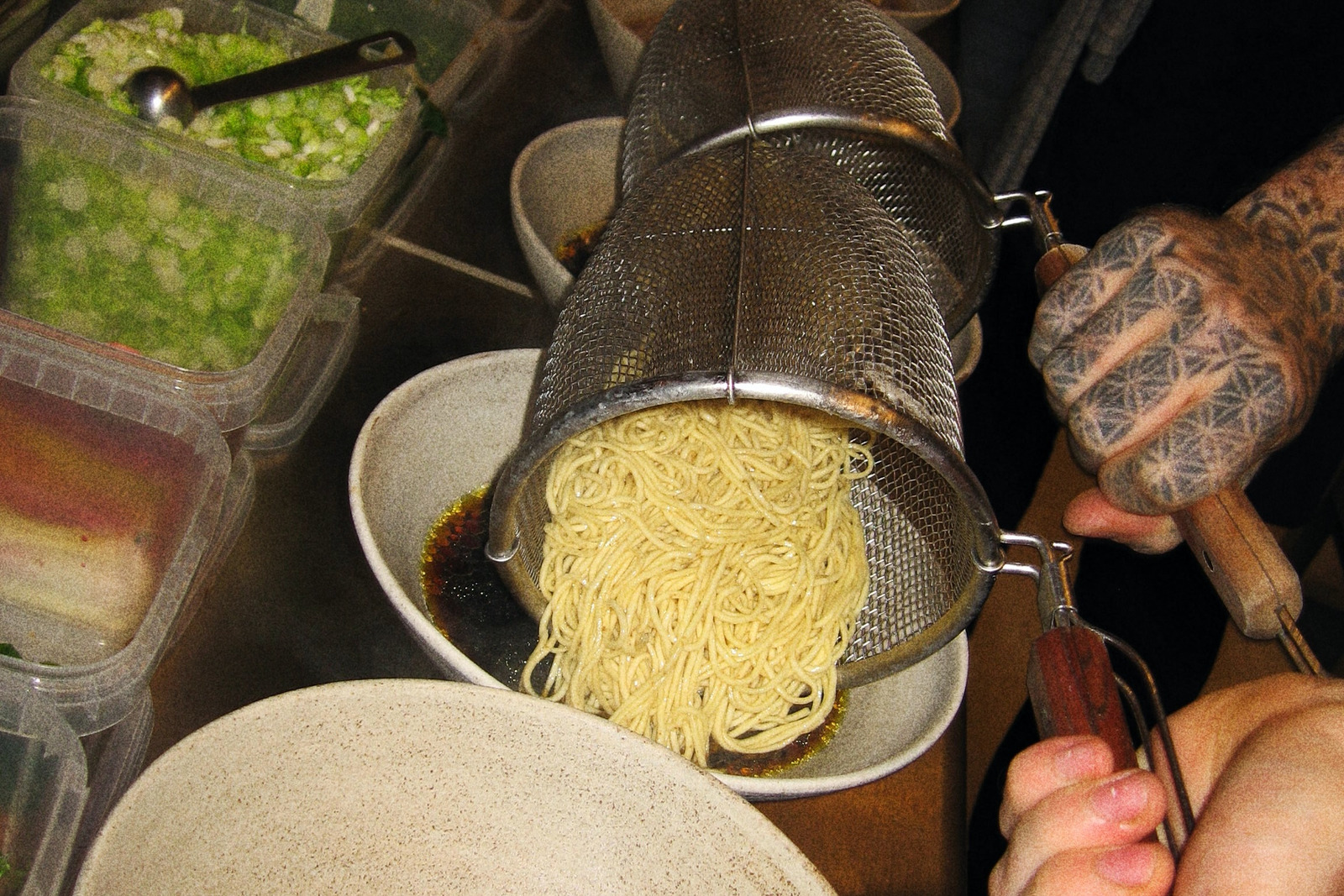 Cooked noodles tipped from a steel mesh basket into a bowl of dark sauce, held by two hands, with tubs of chopped spring onion beside it
