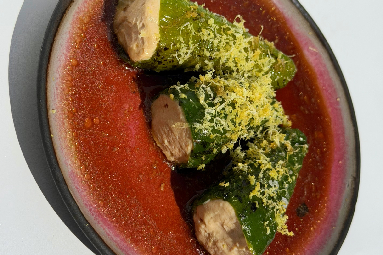 Three tofu-stuffed padrón peppers topped with grated lemon zest in a red sauce on a round plate