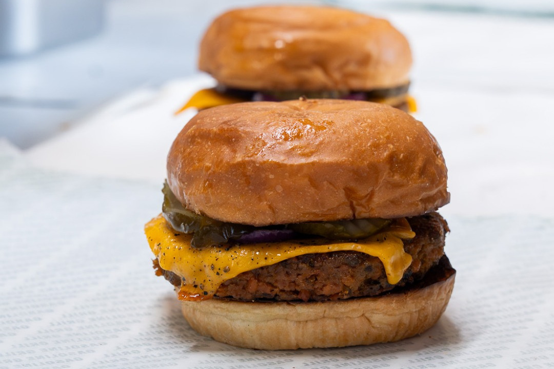 A cheeseburger with a Matr plant patty, melted cheddar, pickles and red onion in a glazed bun, with a second burger behind it
