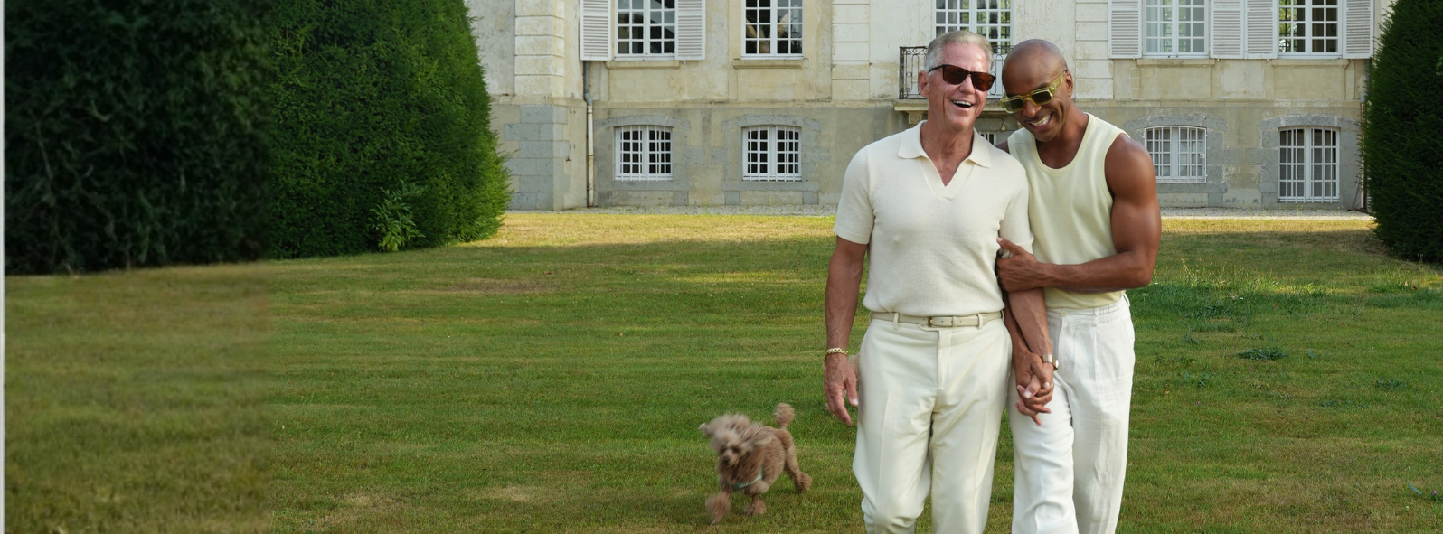 Two men smiling and holding hands, walking on grass with a small dog running nearby by a large stone building.