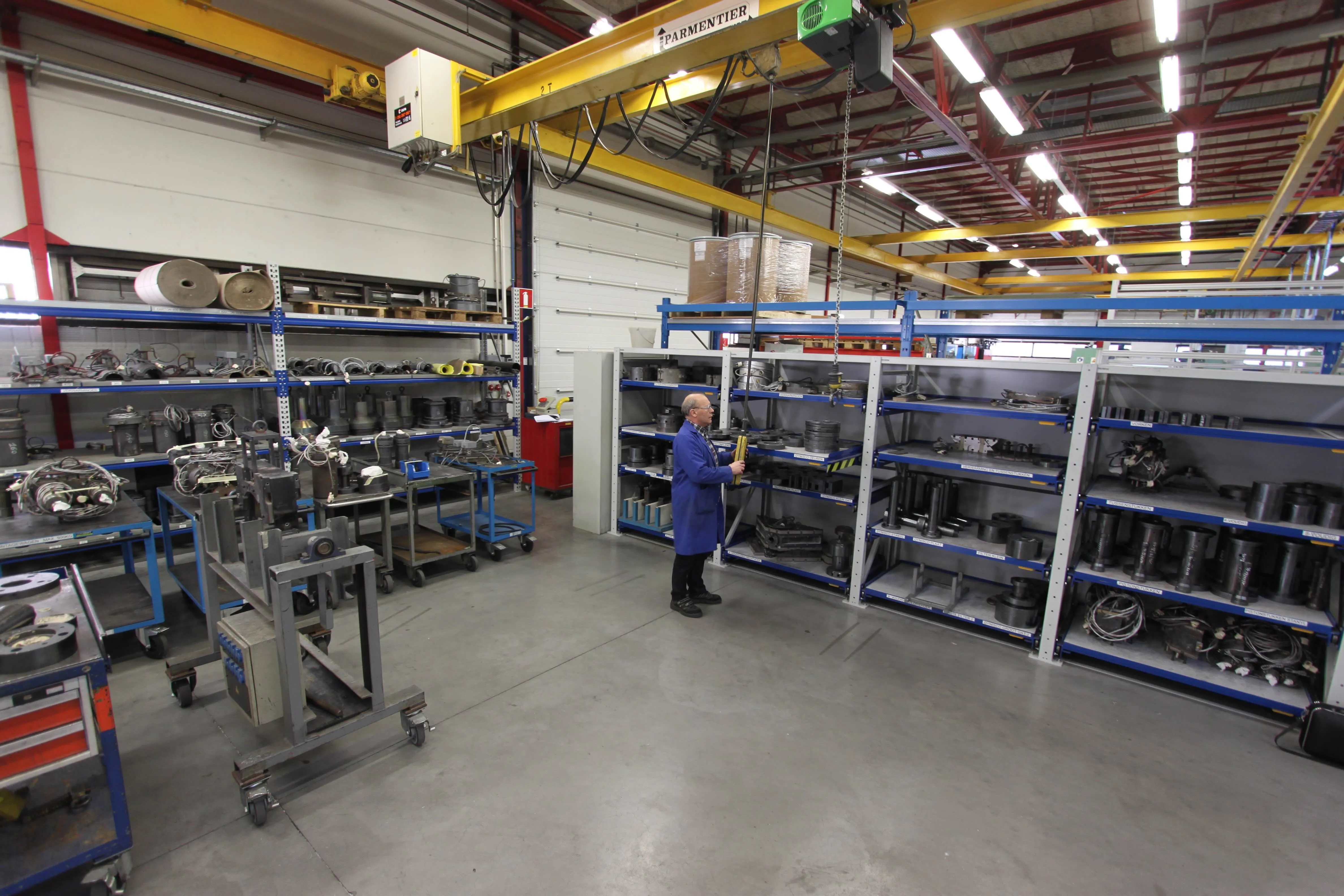 Man in blauwe jas bedient hijskraan in magazijn met metalen onderdelen op rekken.