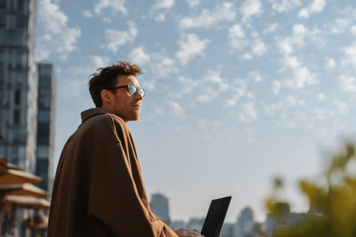 Man wearing sunglasses working on a laptop outdoors with a cityscape and cloudy sky in the background.
