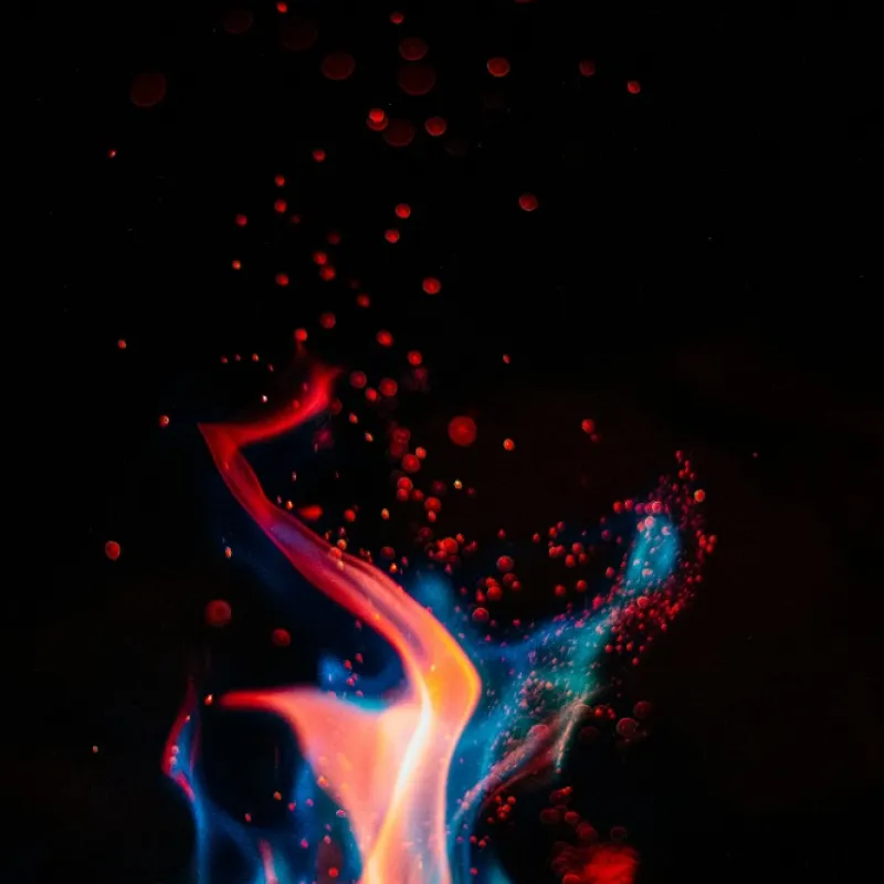 Abstract, colorful flame with blue and orange hues surrounded by scattered red particles against a black background.