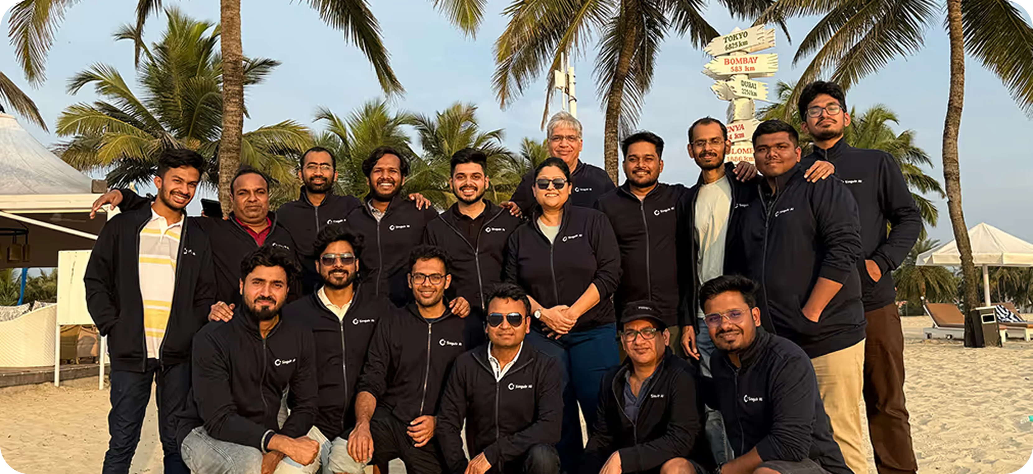 Group of sixteen people in matching black jackets posing and smiling on a sandy beach with palm trees and signpost in the background.