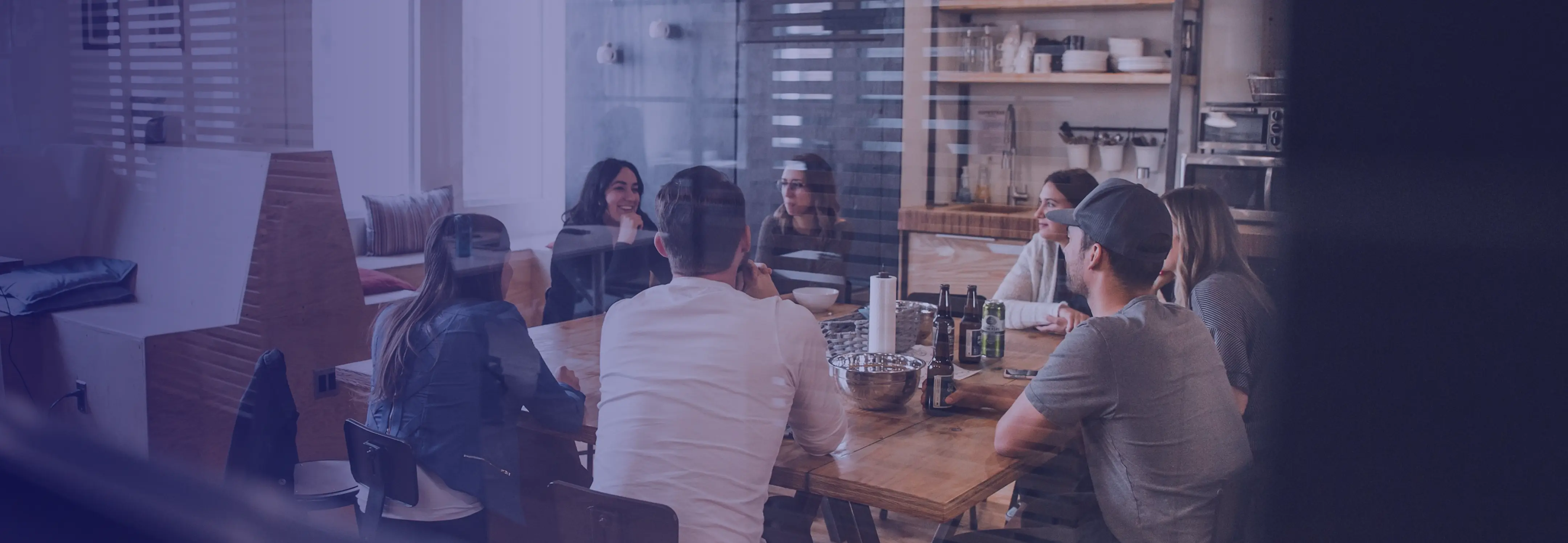 Group of people having a meeting around a wooden table with drinks and bowls in a modern office kitchen.