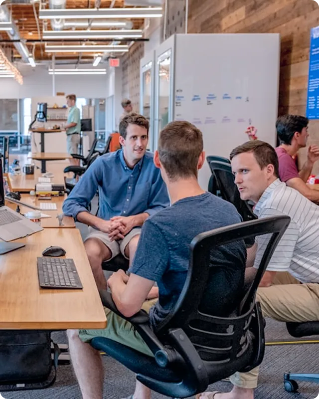 Three men engaged in a discussion in an open office space with computers and modern office furniture.
