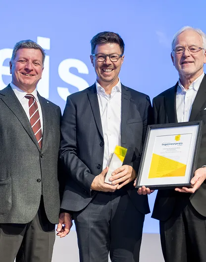 Drei Männer in Anzügen lächeln, einer hält eine Auszeichnung, ein anderer ein gerahmtes Zertifikat bei einem Preisverleihungsevent.