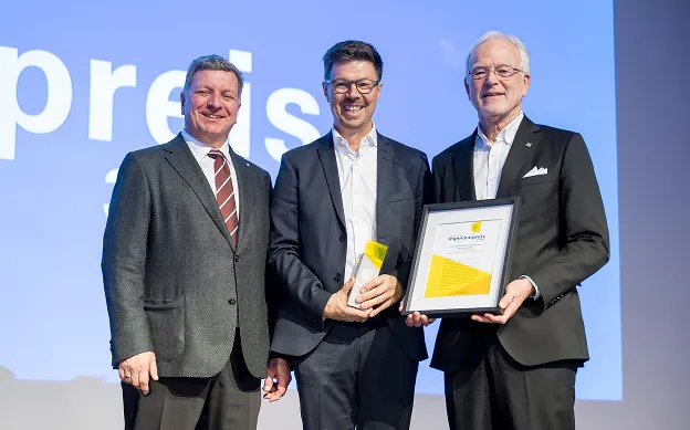 Drei Männer in Anzügen posieren mit einem Award und einer Urkunde vor einem blauen Hintergrund.