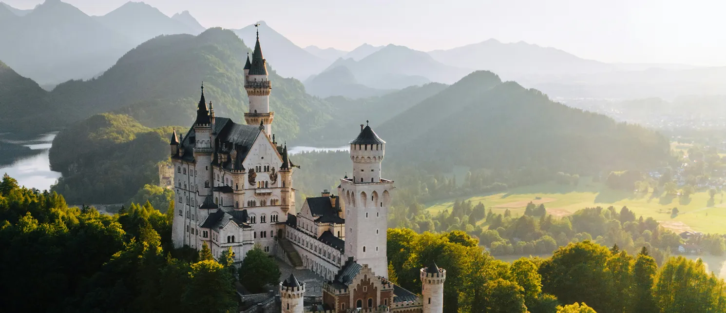 Das Schloss Neuschwanstein in Bayern, eingebettet in grüne Wälder mit Bergen und einem See im Hintergrund bei Sonnenlicht.
