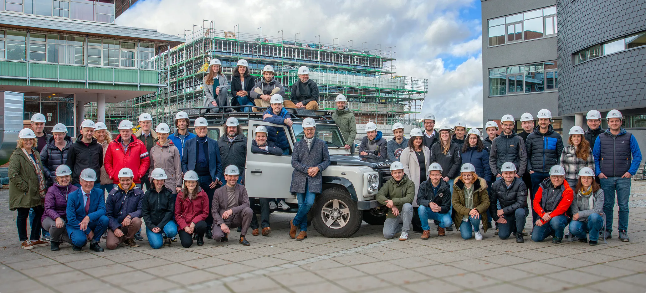 Gruppenfoto von 40 Personen mit weißen Bauhelmen, die vor einem grauen Geländewagen und einem Gebäude mit Baugerüsten stehen.