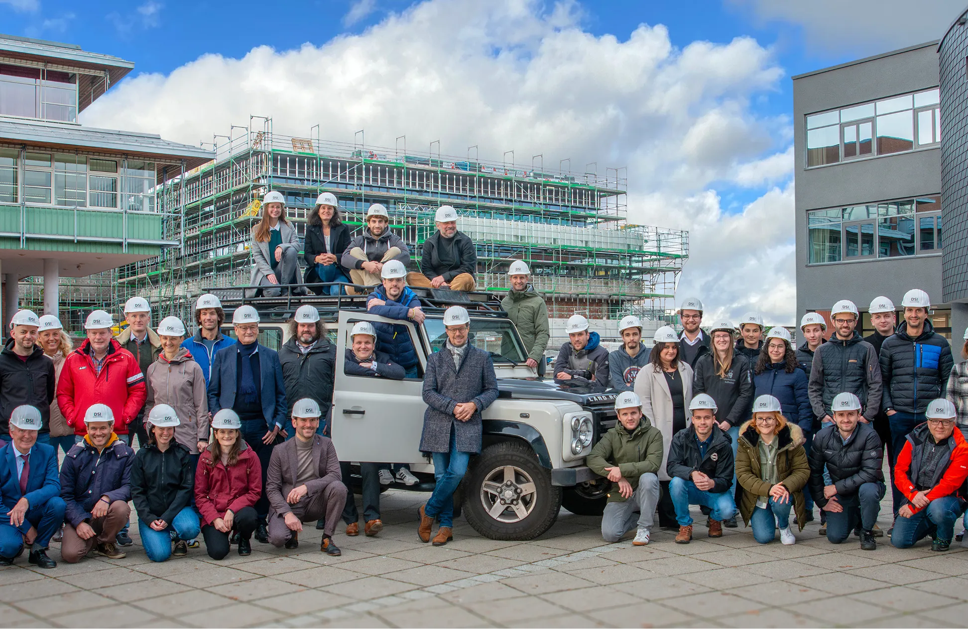 Eine große Gruppe von Menschen mit weißen Helmen steht und kniet um einen weißen Land Rover vor einer Baustelle mit einem mit Gerüsten versehenen Gebäude unter einem bewölkten Himmel.