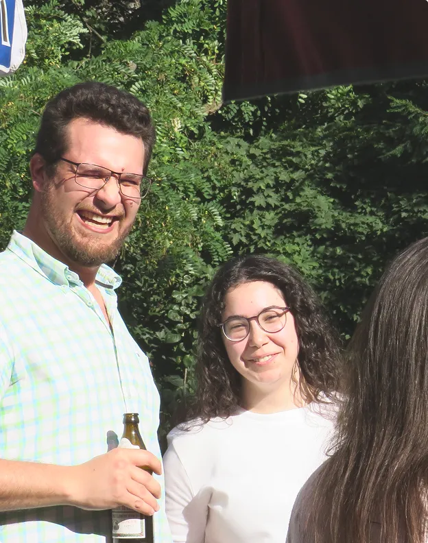Mann mit Brille und kariertem Hemd hält eine Flasche, neben einer lächelnden Frau mit Brille und langem lockigem Haar, im Hintergrund grüne Büsche.