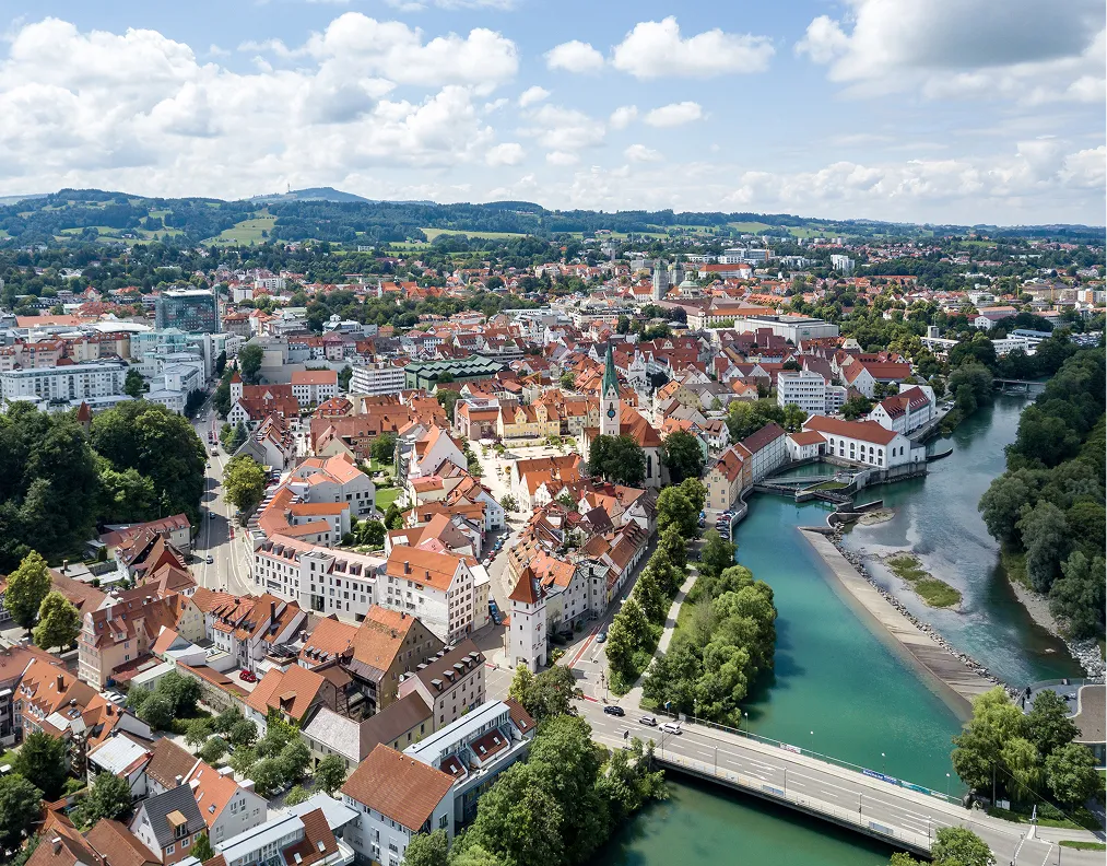 Luftaufnahme der Stadt Kempten mit roten Dächern, Fluss und bewaldeten Hügeln im Hintergrund bei sonnigem Himmel.
